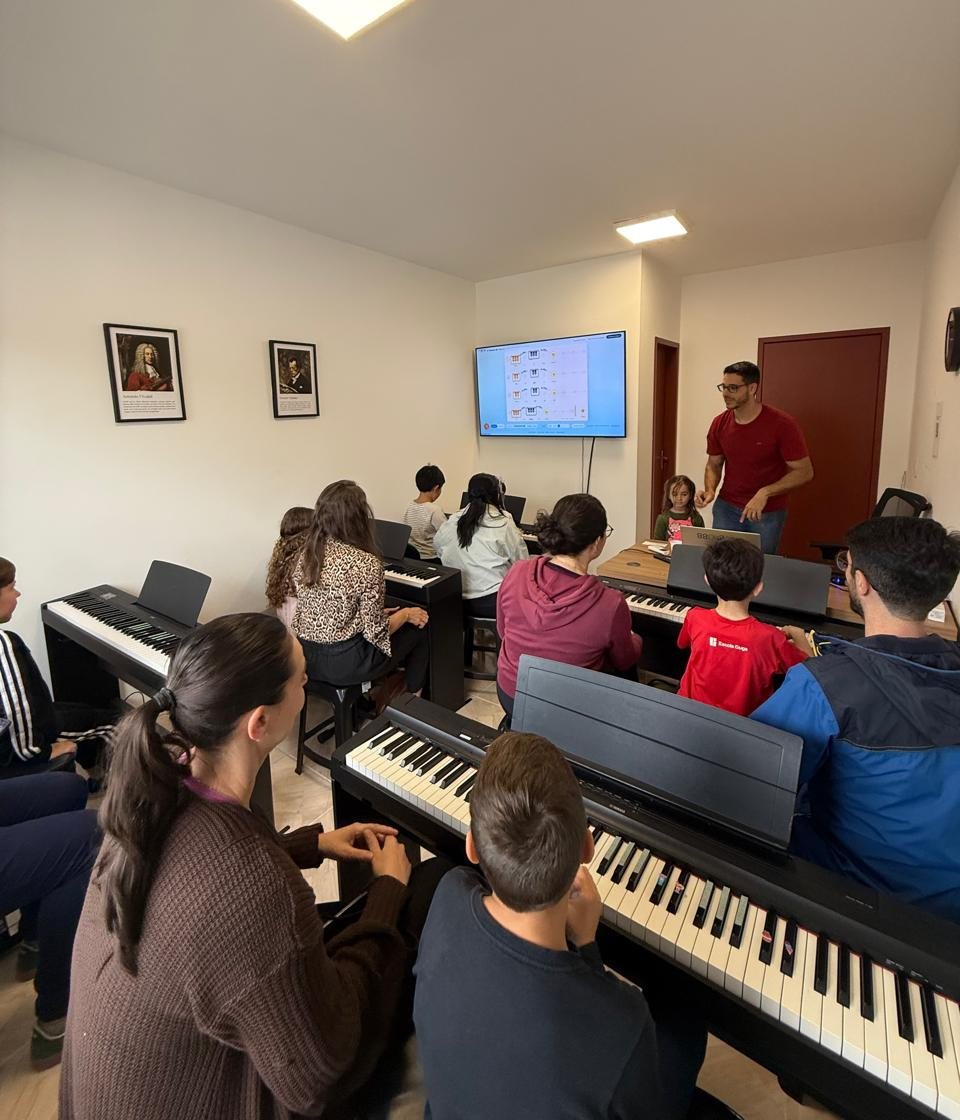 Aula de Piano Kids em grupo no Studio88: crianças em pianos digitais individuais, pais sentados ao lado e o jogo da aula na tela da sala